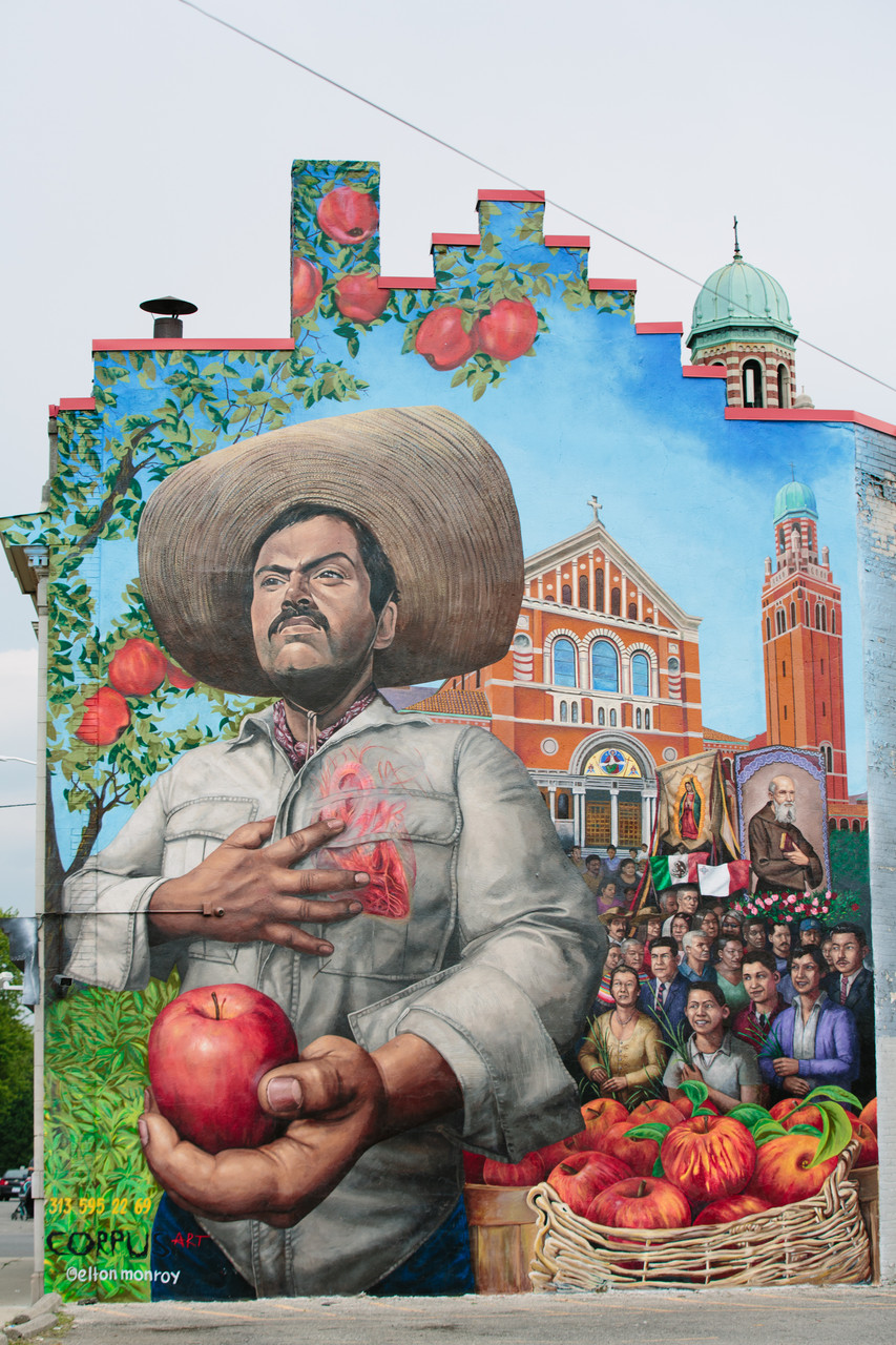 A mural depicting a Mexican man in the foreground holding an apple in one hand and the other hand over his chest, facing away from a community of people holding flags and tapestries with a church visible in the distance.